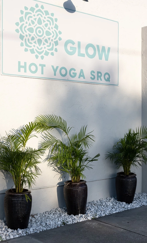 Exterior sign for Glow Hot Yoga SRQ in Sarasota with three potted palm plants along the building entrance