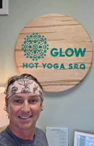 Instructor wearing a headband stands in front of a round wooden sign that reads "GLOW HOT YOGA SRQ" with a turquoise mandala design.